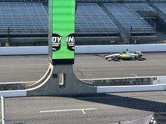 Green and white Formula racing car driving on a racetrack with empty grandstands in the background and a green vertical banner displaying 'INDY' on a pole.