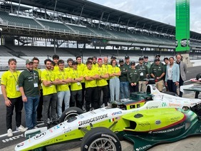 MSU engineering team standing behind a green and white Formula racing car in the pit area of a professional racetrack, with the grandstands visible in the background.